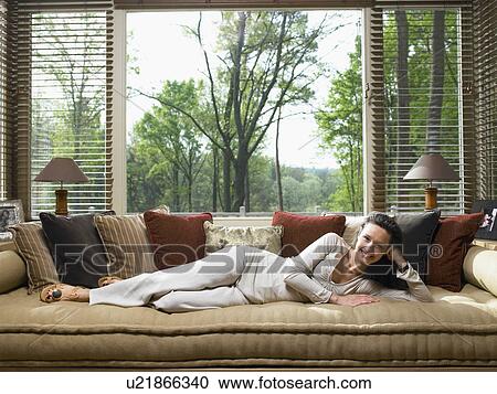 Stock Image - Woman lying on couch in living room smiling.. Fotosearch