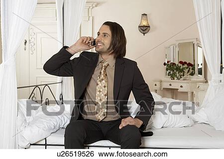 Stock Photograph - young businessman smiling holding mobile phone sitting on bed. Fotosearch