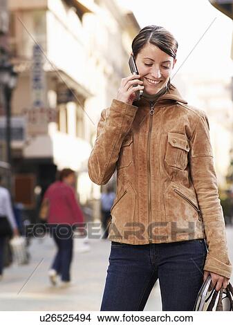 Picture - Young woman standing in street using mobile phone, smiling. Fotosearch