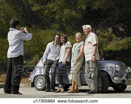 Picture - Multi-generational family standing beside car, boy (8-10) taking photo. Fotosearch