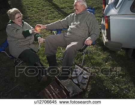 Stock Photography - Senior couple having picnic by car, holding apple, smiling. Fotosearch