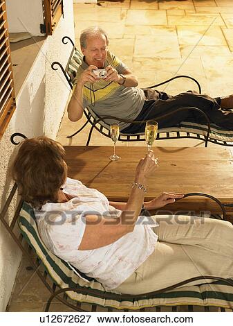 Senior couple lying on sun loungers, man photographing woman raising glass of champagne View Large Photo Image Stock Photo - Senior couple lying on sun loungers, man photographing woman raising glass of champagne. Fotosearch