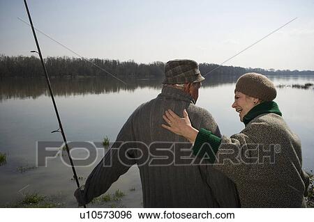 Stock Photograph - Senior couple standing by river, man holding fishing rod, rear view. Fotosearch