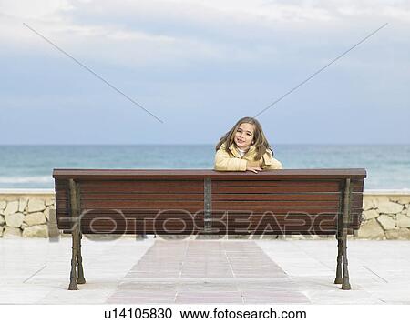 Stock Image - Little girl (6-8) looking over the back of a bench, sea in background. Alicante, Spain.. Fotosearch