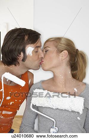 Stock Image - Young couple holding paint rollers, kissing. Fotosearch