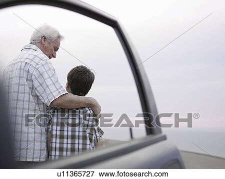 Stock Photo - Boy (8-10) and grandfather standing beside car at beach, rear view. Fotosearch