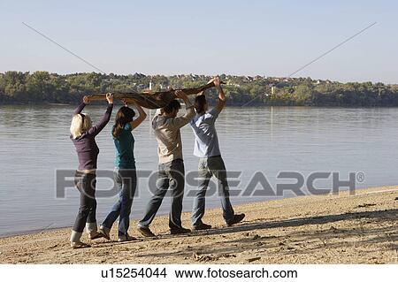 Picture - Four friends carrying a log by a lake.. Fotosearch