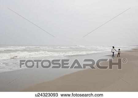 Stock Image - Mother and daughter (9-11) walking along beach, rear view. Fotosearch