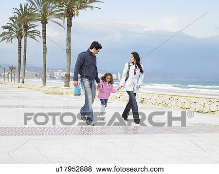 Stock Photo - Mother and father walking along palm lined pavement by the sea with young daughter (6-8). Alicante, Spain.. Fotosearch