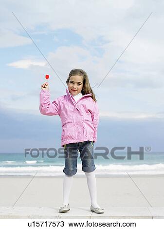 Stock Image - Young girl (6-8) holding lollipop in front of sea, portrait. Alicante, Spain.. Fotosearch