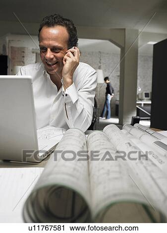 Stock Image - Architect talking on phone while using laptop, female in background. Brussels, Belgium.. Fotosearch