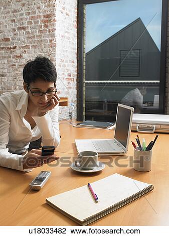 Stock Image - Businesswoman at desk reading messages on her mobile. Brussels, Belgium.. Fotosearch