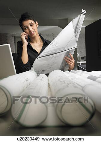 Stock Photo - Female architect talking on the phone while reading blueprint at desk. Brussels, Belgium.. Fotosearch