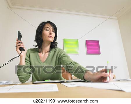 Stock Photo - Businesswoman writing and giving a call at her desk.. Fotosearch