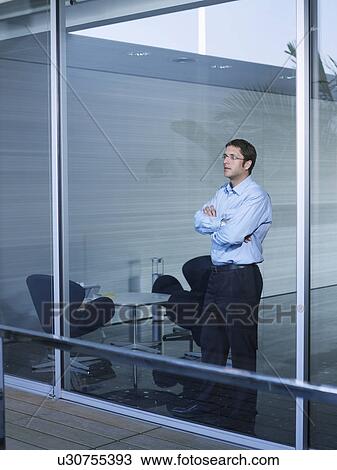 Stock Image - Young businessman standing at window in office, arms crossed. Fotosearch