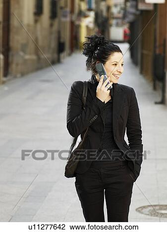 Stock Image - Young woman walking in street using mobile phone, smiling. Fotosearch