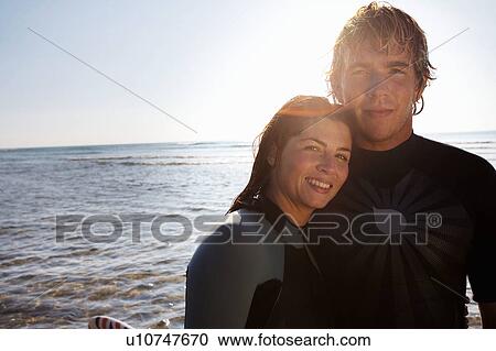 Stock Image - Couple standing on beach with surfboard.. Fotosearch