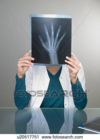 Stock Image - Female doctor holding x ray of hand in front of her head at her desk.. Fotosearch