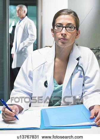 Stock Image - Portrait of female doctor at her desk with male doctor in background.. Fotosearch