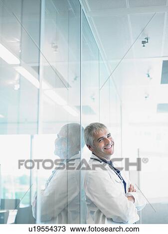 Smiling male doctor leaning on glass wall in hospital lobby. View Large Photo Image Stock Image - Smiling male doctor leaning on glass wall in hospital lobby.. Fotosearch