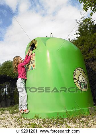 Stock Image - Side view of young girl (5-7) putting glass bottle in recycling bin, Alicante, Spain,. Fotosearch