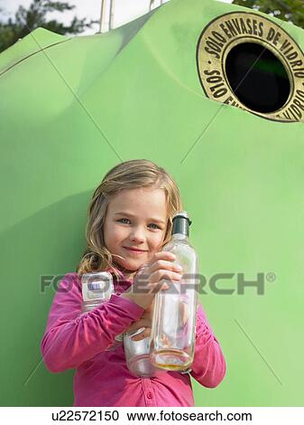 Stock Image - Young girl (5-7) standing in front of recycling bin holding glass bottles, Alicante, Spain,. Fotosearch