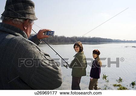 Stock Photo - Grandfather photographing grandson (12-14) and granddaughter (10-12) fishing in river. Fotosearch