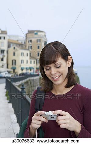 Stock Image - Young woman portrait with camera looking at display smiling old city in background.. Fotosearch