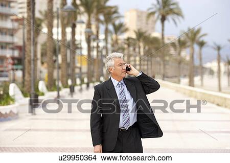 Picture - Senior businessman talking on mobile phone whilst walking along palm lined pavement. Alicante, Spain.. Fotosearch