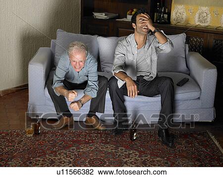 Stock Image - Senior father and son sitting on sofa watching television. Fotosearch