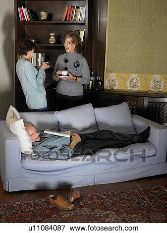 Stock Photo - Senior man asleep on sofa, two women chatting in background. Fotosearch