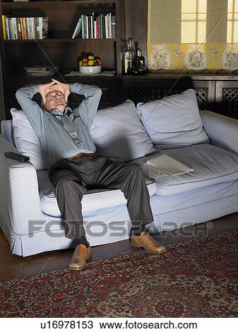 Stock Image - Senior man holding forehead sitting on sofa illuminated by television. Fotosearch