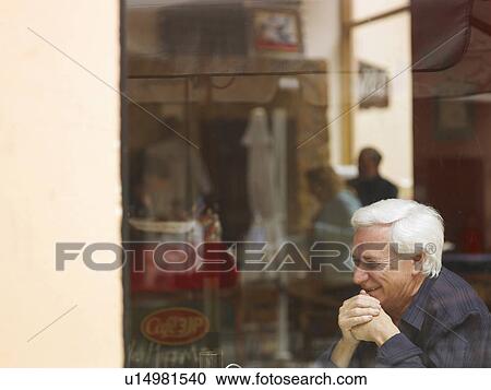 Stock Image - Senior man sitting at café table, laughing, view through window, side view. Fotosearch