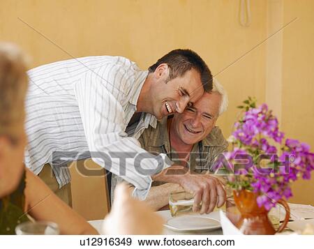 Stock Photo - Senior man sitting at dining table on balcony, adult son pouring wine. Fotosearch
