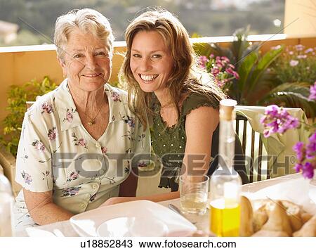 Stock Image - Senior woman and adult daughter sitting at dining table on balcony, portrait. Fotosearch