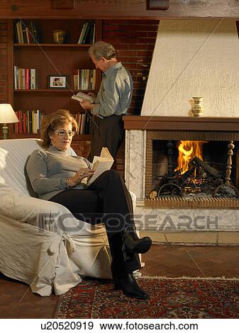 Stock Photo - Senior woman reading book by fire in living room, man in background. Fotosearch
