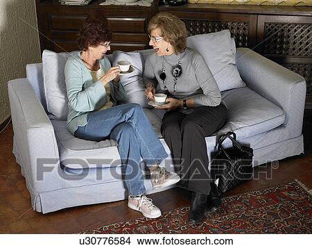 Two senior women sitting on sofa holding cups, laughing View Large Photo Image Picture - Two senior women sitting on sofa holding cups, laughing. Fotosearch