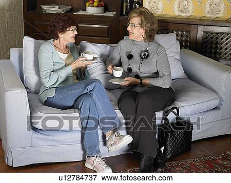 Two senior women sitting on sofa holding cups, smiling View Large Photo Image Stock Photo - Two senior women sitting on sofa holding cups, smiling. Fotosearch