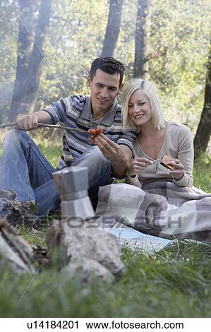 Stock Image - Couple at campsite cooking hot dogs and smiling.. Fotosearch