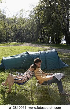 Stock Image - Couple sitting back to back at campsite with the man reading a newspaper.. Fotosearch
