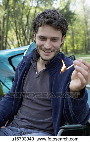 Stock Photo - Man sitting at campsite holding a lit match smiling.. Fotosearch