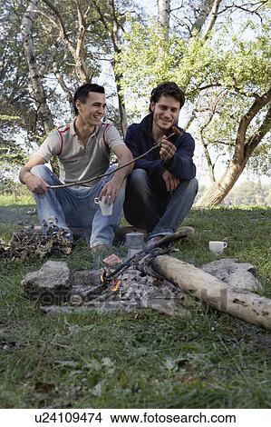 Picture - Two men cooking hot dogs over a fire pit smiling.. Fotosearch