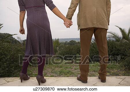 Stock Image - Couple holding hands outside, low section, rear view. Fotosearch
