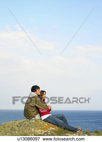 Stock Photo - Couple sitting on a cliff looking out to sea.. Fotosearch
