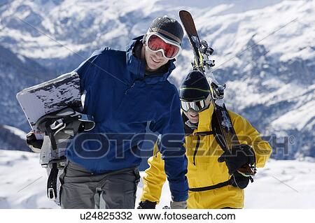 Stock Image - Male skier and male snowboarder walking up mountain ridge carrying skis. Fotosearch
