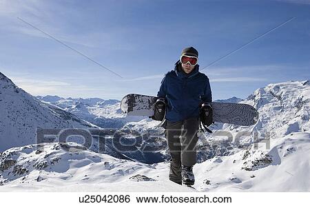 Stock Photograph - Male snowboarder walking up mountain ridge carrying snowboard. Fotosearch