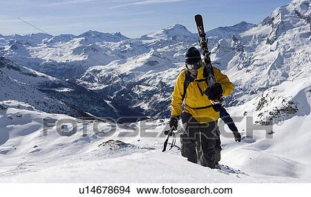 Picture - Two male skiers walking up mountain ridge carrying skis. Fotosearch