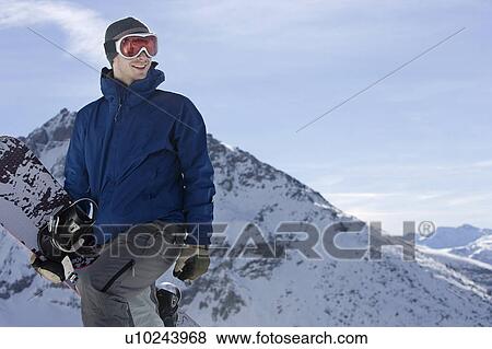 Stock Photo - Male snowboarder standing on top of mountain ridge with snowboard. Fotosearch