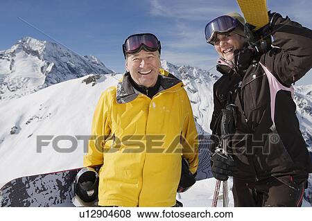 Stock Photo - Mature couple in ski-wear holding Skis and snowboard on mountain. Fotosearch