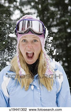 Stock Image - Snowball exploding on young blonde woman wearing ski-wear in forest. Fotosearch
